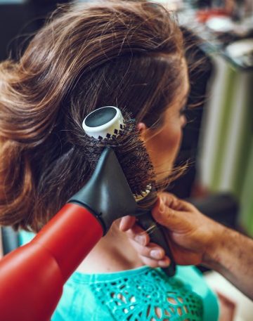 Male Hairdresser working in a beauty salon and drying brown hair with round brush of a female customer.