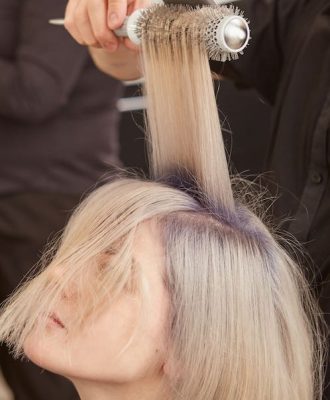 Skillful male hairstylist doing blowout hairstyle for blonde young woman in beauty salon